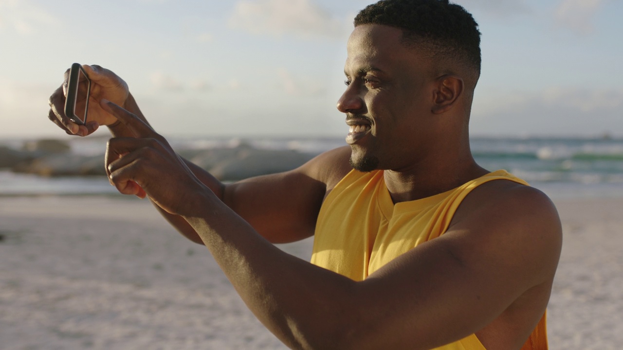 年轻健康的非洲裔美国人穿着黄色背心在海滩上用智能手机拍照视频素材