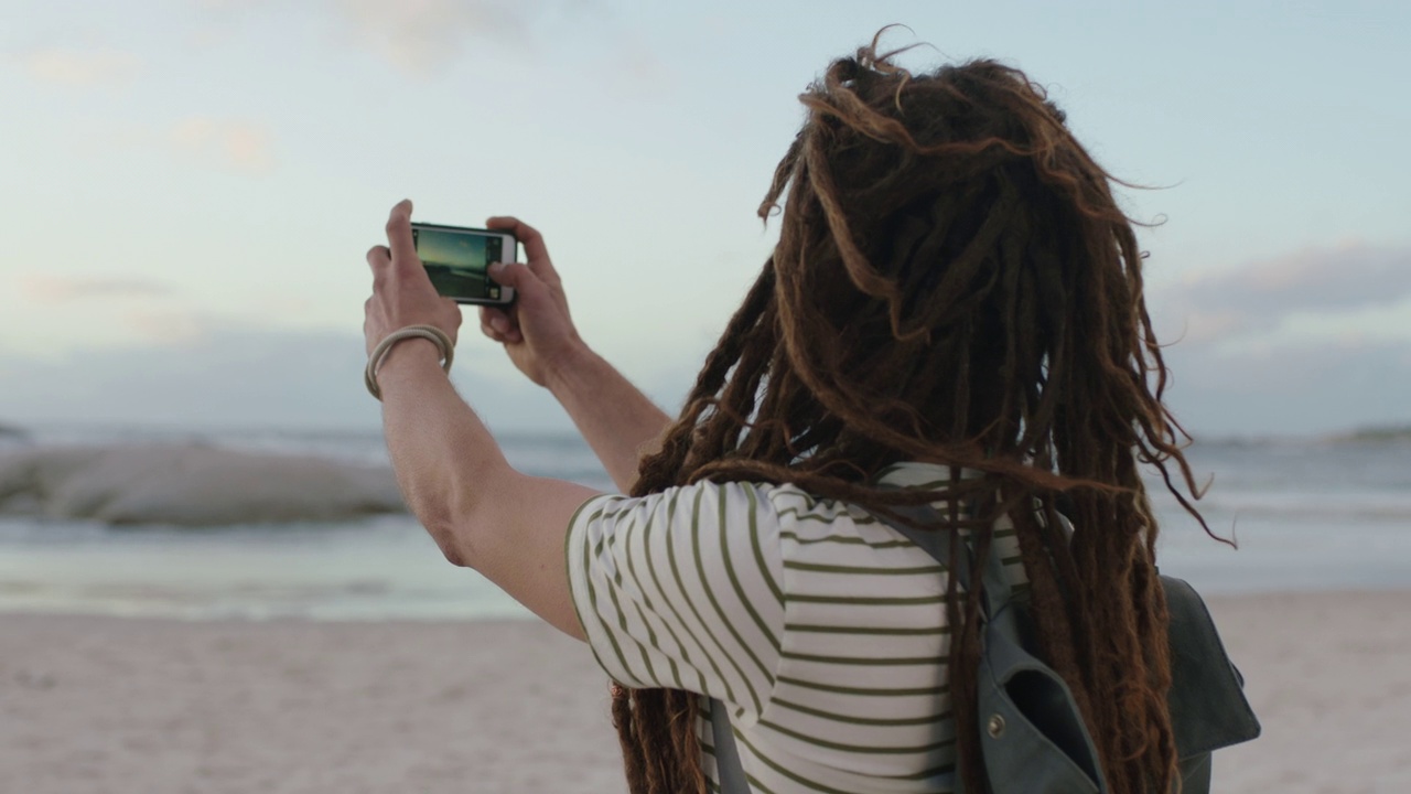 留着脏辫的无忧无虑的年轻人穿着条纹衬衫在海滩上用手机拍照视频素材