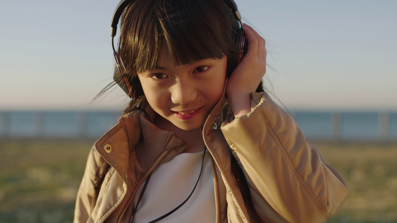 可爱的亚洲小女孩戴着耳机，在日落的海边海滩公园微笑着听音乐视频素材