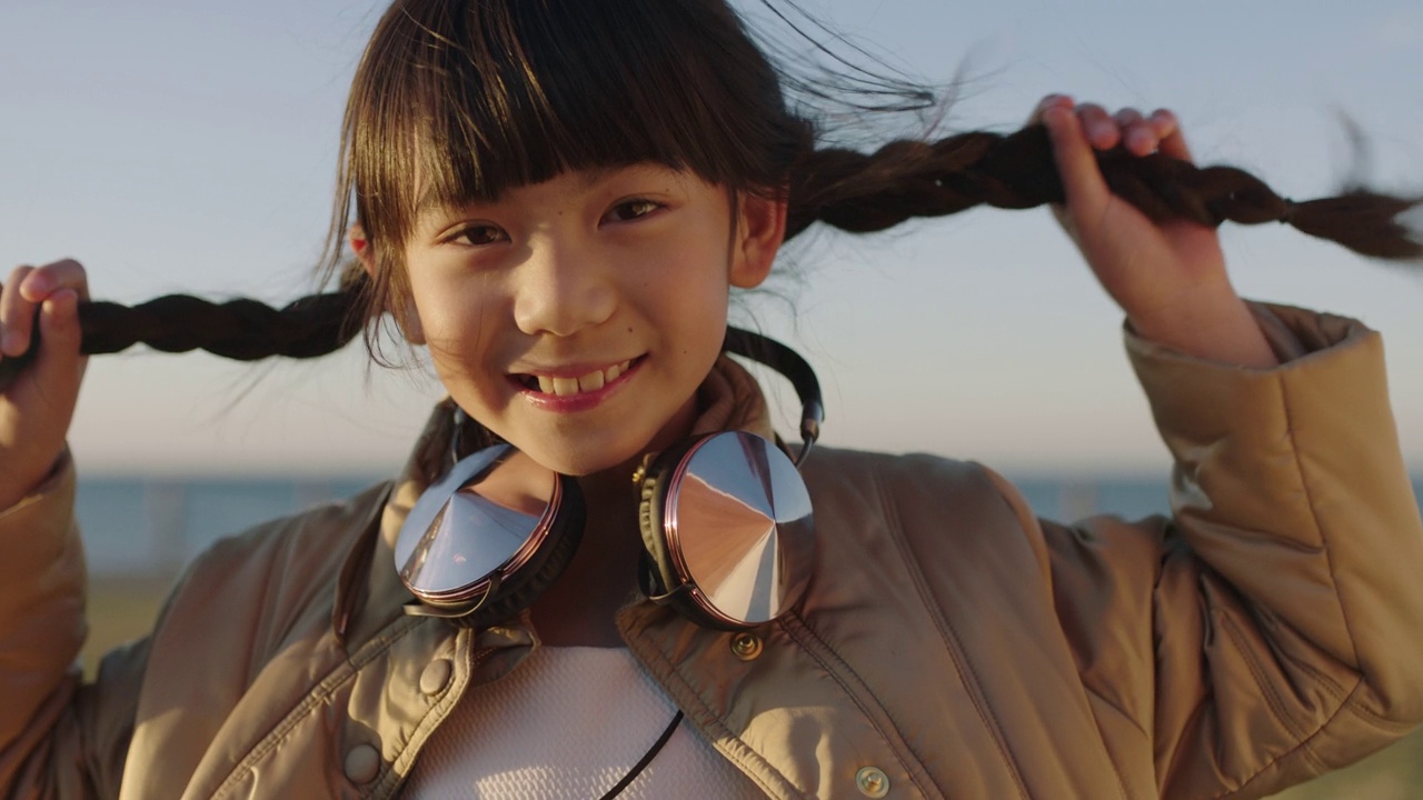 欢快的亚洲女孩的特写肖像，微笑着，戴着耳机，在海边海滩公园享受快乐好玩的一天视频素材