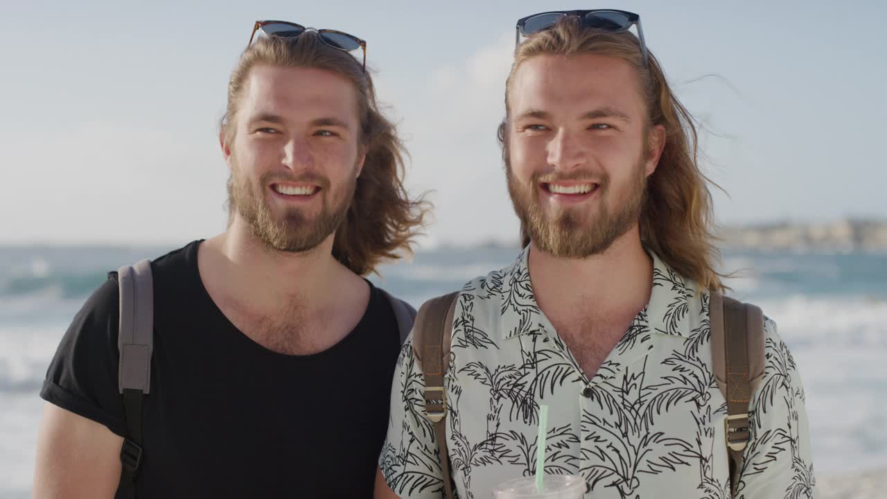英俊的双胞胎兄弟在海滩上微笑，在海边享受暑假，喝着果汁饮料视频下载