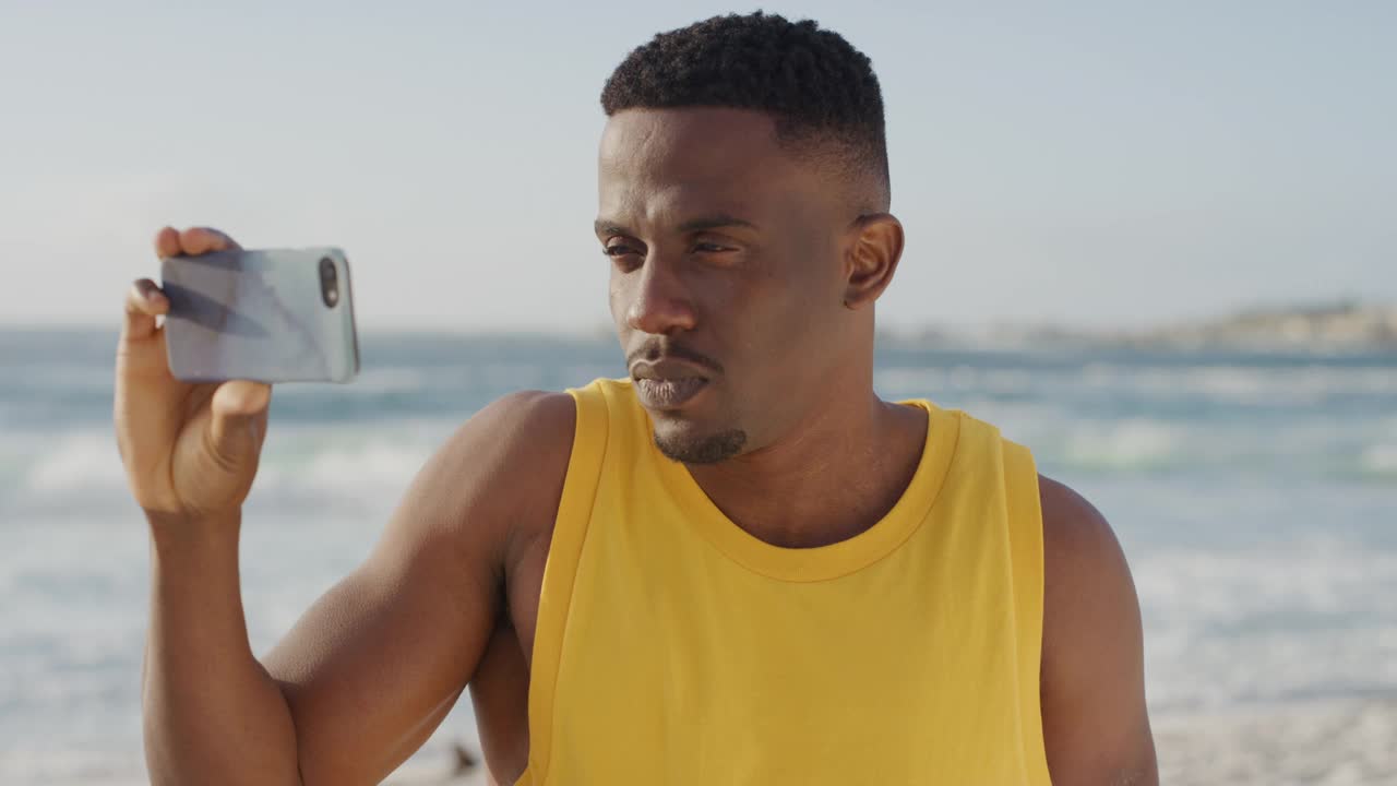 年轻英俊的非洲裔美国男子穿着黄色背心，在海滩度假时用智能手机拍照，微笑着享受移动技术视频下载