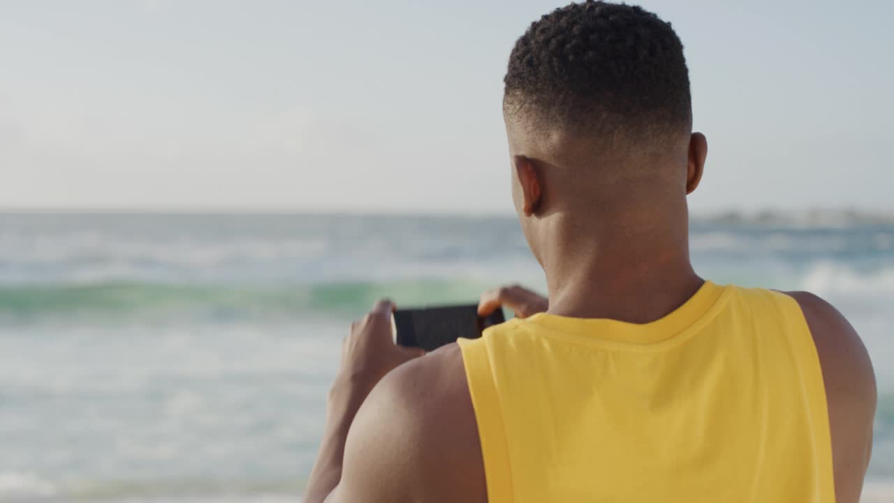 年轻的肌肉发达的非洲裔美国人用智能手机拍摄美丽的海洋海滨，穿着黄色背心在海滩上享受度假手机分享视频素材