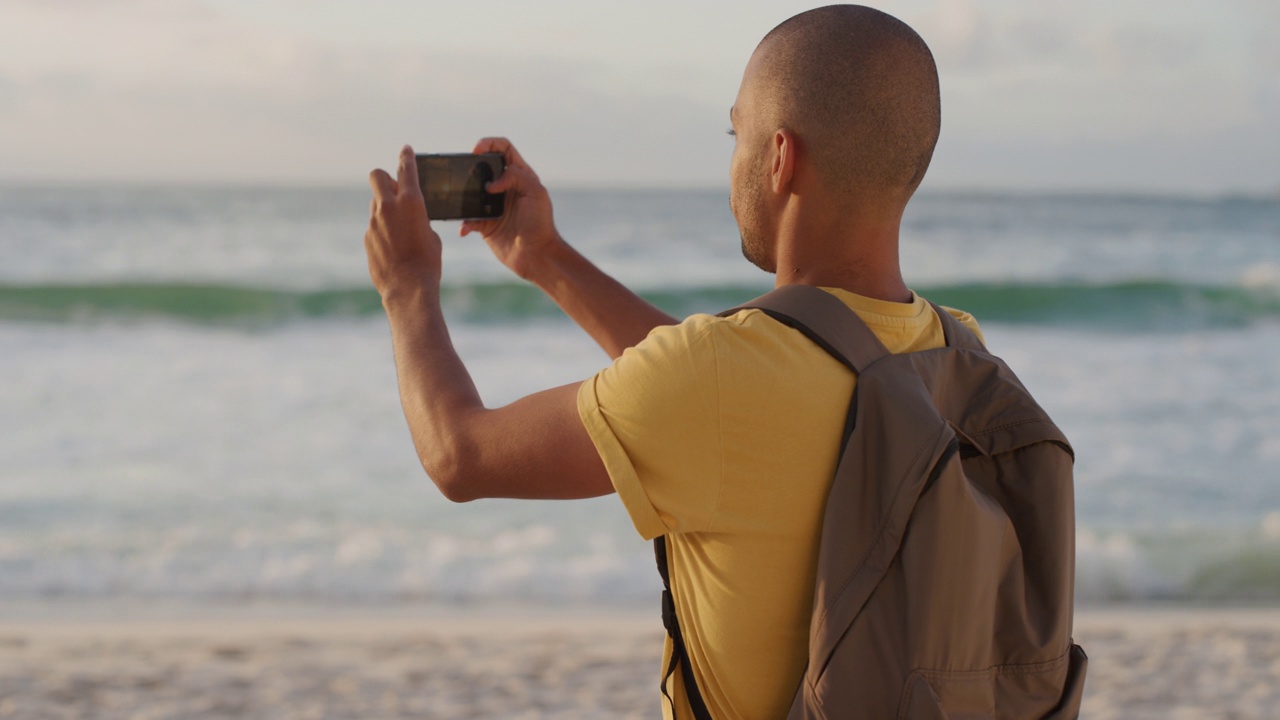 年轻的西班牙裔男子用智能手机在海滩上拍摄美丽的海洋海滨，享受美丽的日落景色，分享度假体验视频素材