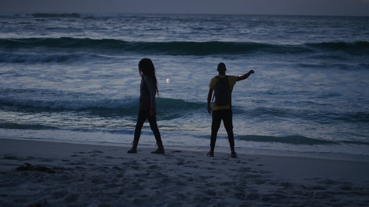 欢快的朋友们在沙滩上跳舞庆祝新年前夜，挥舞着烟花，在日落的平静的海边背景下一起享受愚蠢的乐趣视频下载
