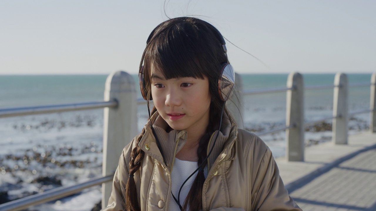 肖像美丽的小亚洲女孩看起来严肃冷静的小孩戴着耳机听音乐在阳光明媚的海边沙滩慢动作视频素材