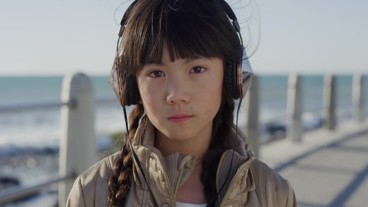 特写肖像漂亮的小亚洲女孩看起来严肃冷静的小孩戴着耳机听音乐在阳光明媚的海边沙滩慢动作视频素材