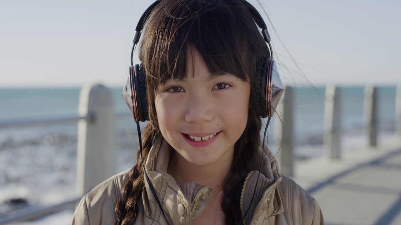 可爱的亚洲小女孩戴着耳机，微笑着在海边的沙滩上听音乐视频素材