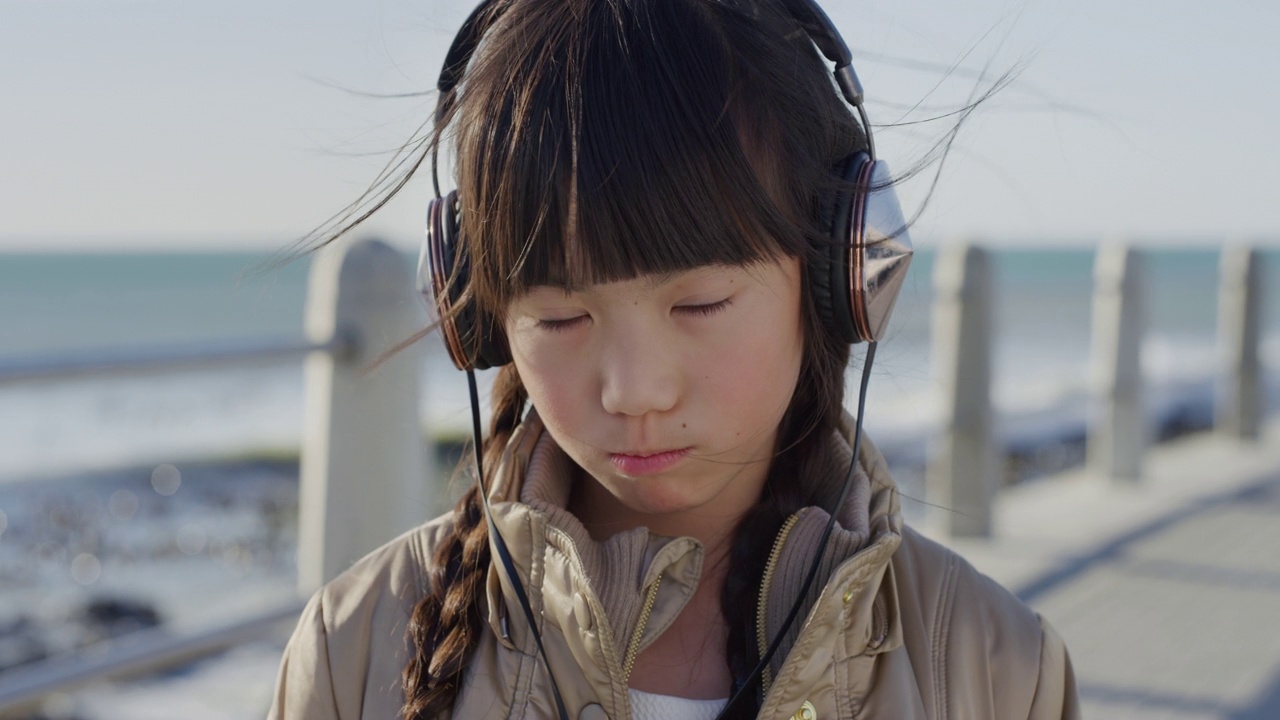 特写肖像美丽的小亚洲女孩看起来生气的小孩心烦戴着耳机在阳光明媚的海边海滩慢动作视频下载