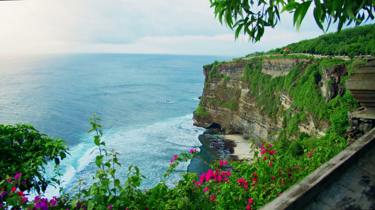 乌鲁瓦图寺庙在高悬崖的边缘俯瞰海水在巴厘岛印度尼西亚。著名的旅游胜地。鲜艳的粉红色花在热带岩石海岸。视频素材