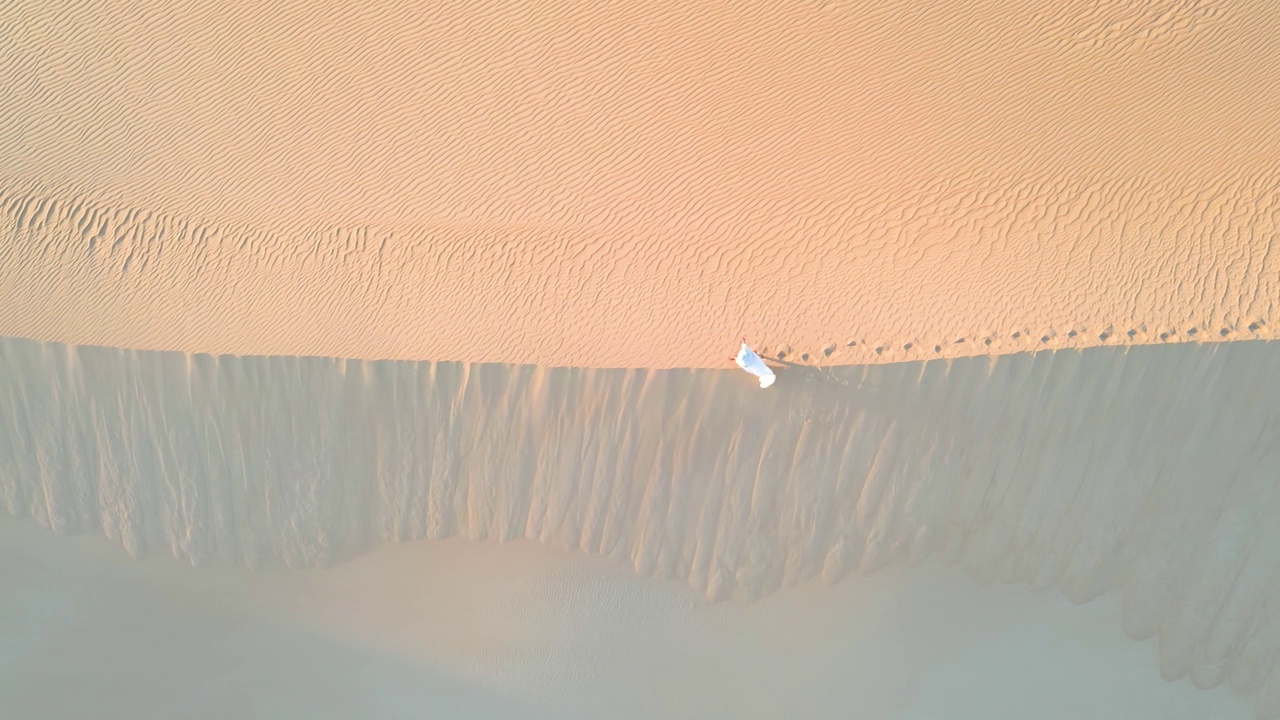 在西部沙漠的黄金时段，一架无人机捕捉到一个迷人的场景，一个女人穿着飘逸的白布优雅地穿过沙丘。视频下载