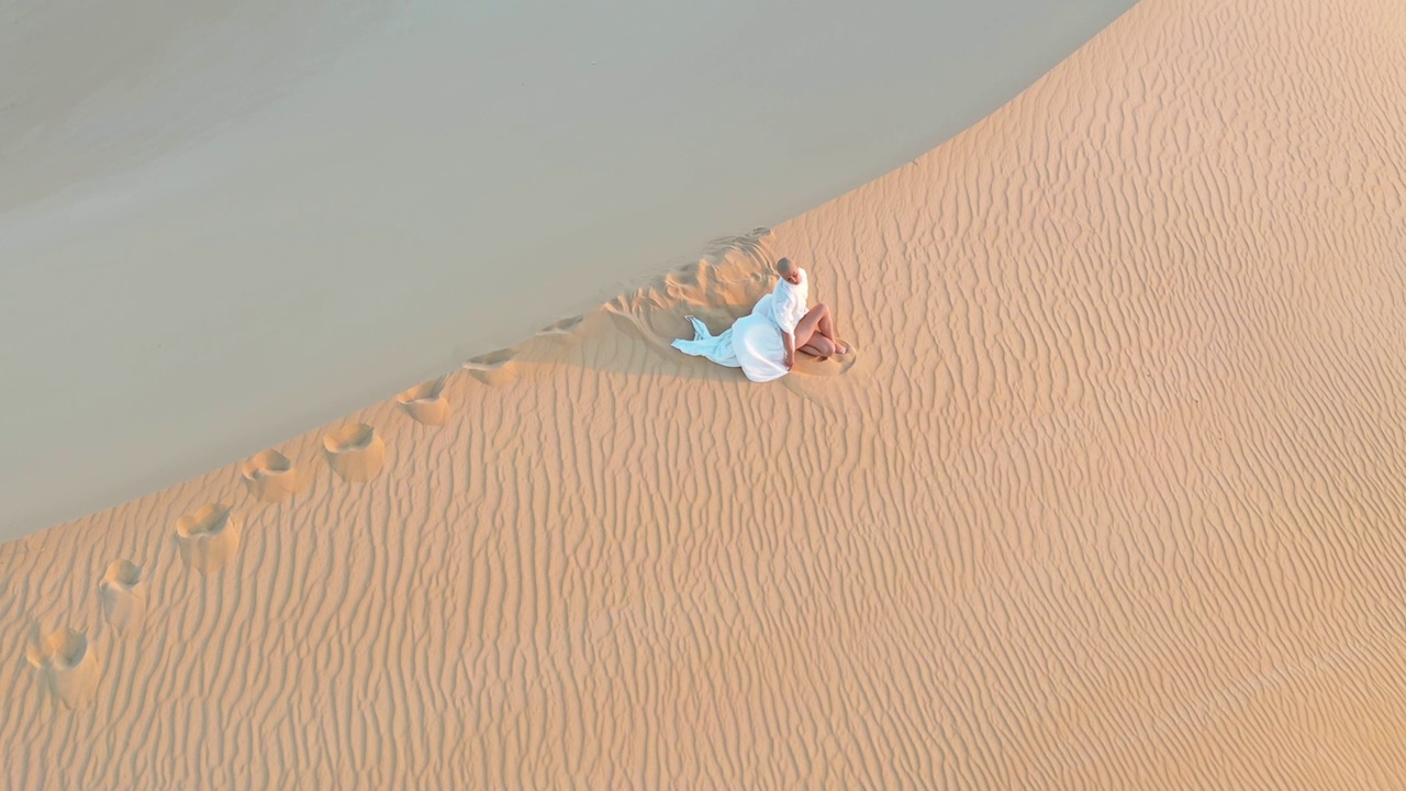航拍视频捕捉到，在黄金时段，一名身穿白色连衣裙的女子坐在埃及撒哈拉沙丘上，创造了一个令人惊叹的场景。视频下载