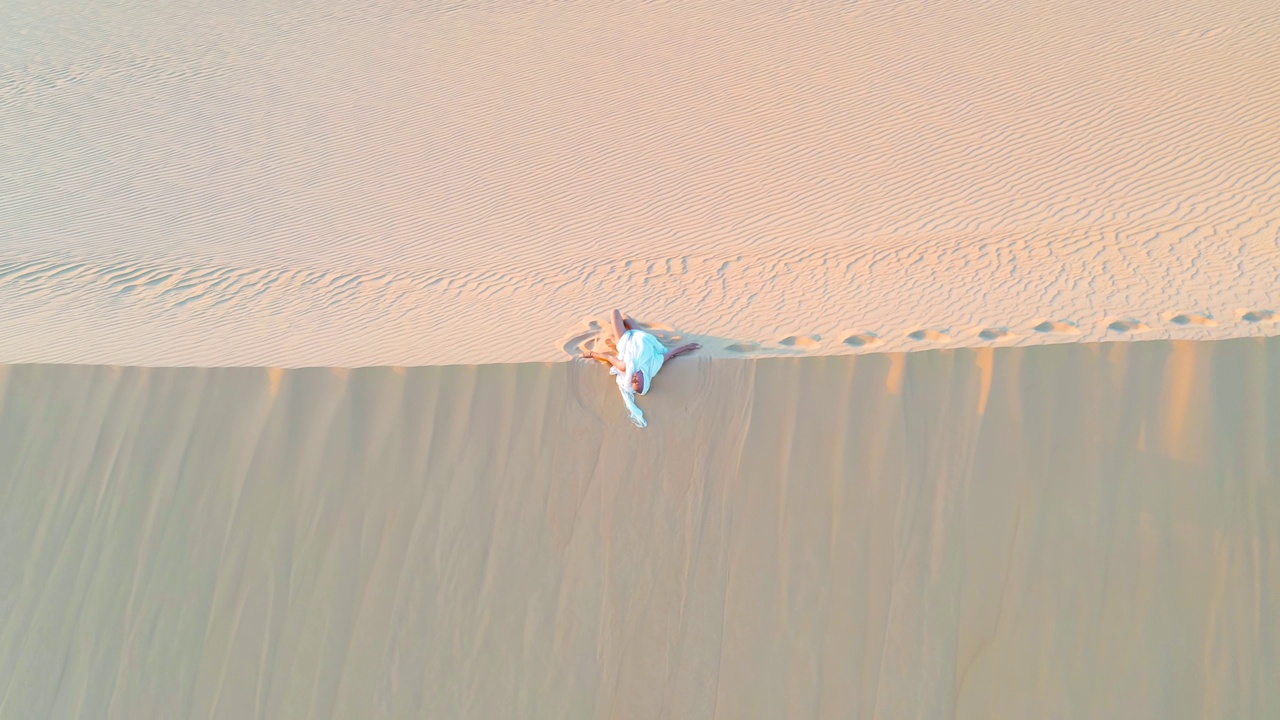 一位身穿白色连衣裙的年轻女子优雅地铲起沙子，让它在风的吹拂下从指尖溜走。视频下载