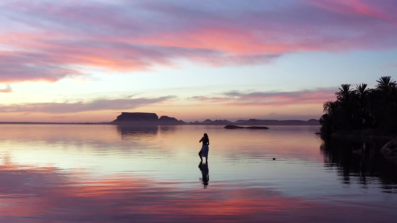 在埃及锡瓦绿洲宁静的氛围中，一个孤独的女人站在浅水里，完全被令人惊叹的日落迷住了。视频下载