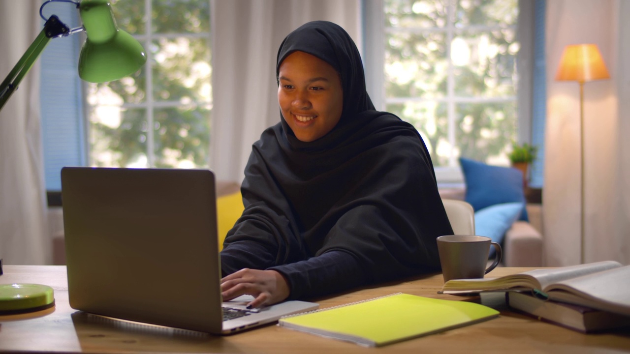 戴着传统头巾的年轻漂亮的穆斯林学生正在用笔记本电脑在家学习。实时视频下载