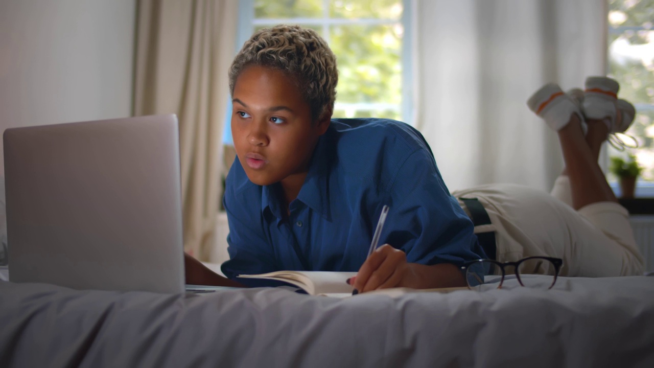 一名非裔美国女学生在床上用笔记本电脑和书本学习。实时视频下载
