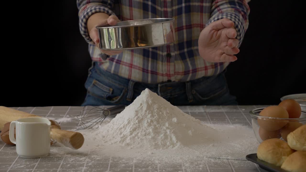手工制作面包面团。用筛子或滤子将面粉筛成一堆面粉，在厨房用烘焙设备工具手工制作面包，黑色背景。专业的烘焙工艺。慢动作视频下载