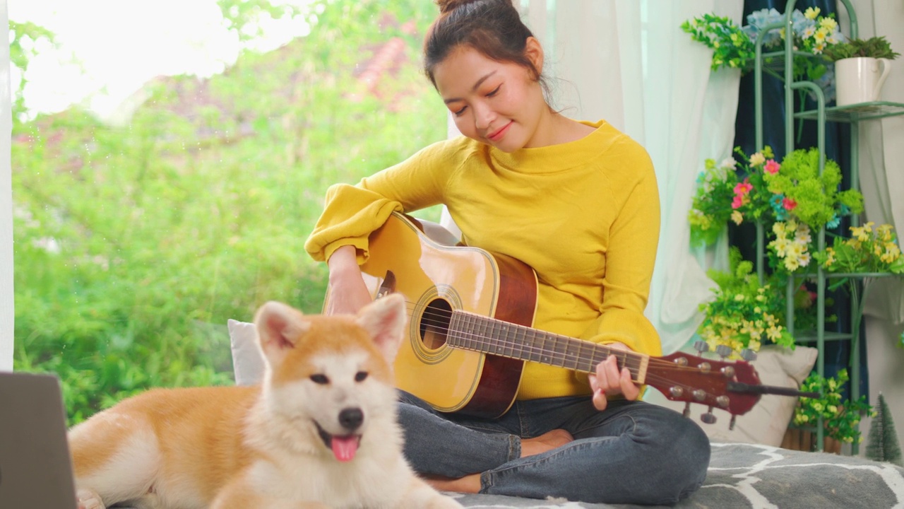 亚洲美女喜欢在家里的客厅里和狗一起弹吉他，宠物看起来很放松，和主人在白天的阳光下呆在一起。视频下载