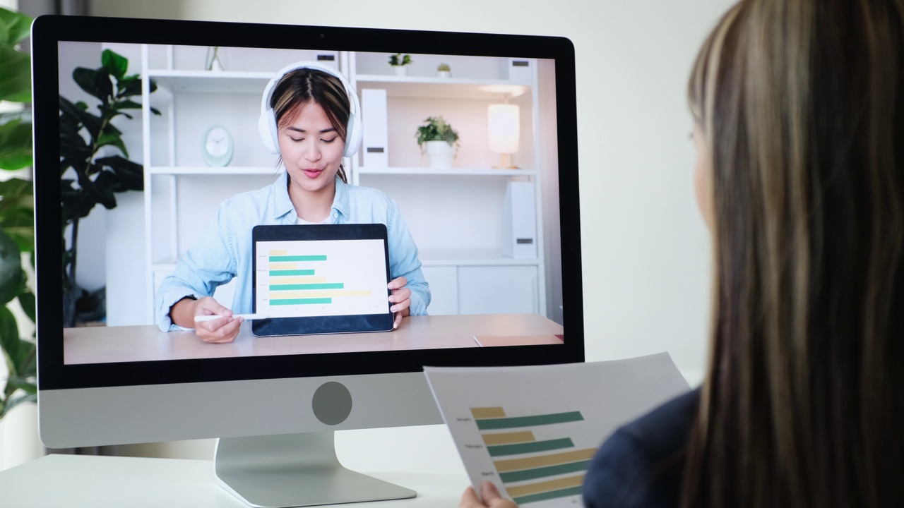 年轻的亚洲女性手持数码平板电脑，在虚拟视频会议中展示销售报告和商业计划，通过家庭办公室的台式电脑与团队进行视频通话在线会议视频下载