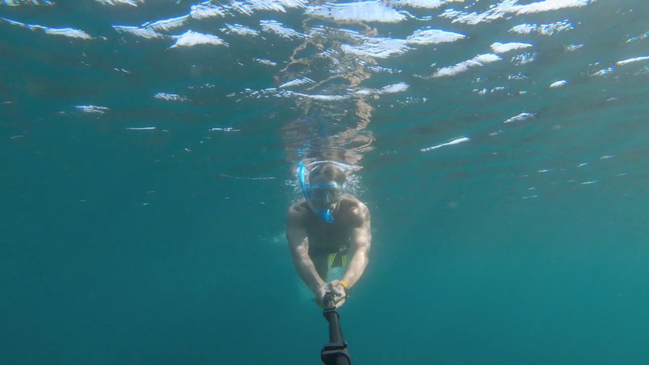 一名白人男子戴着潜水面罩，穿着脚蹼，拿着一根长自拍杆上的运动相机在水下潜水和游泳。浮潜年成视频素材