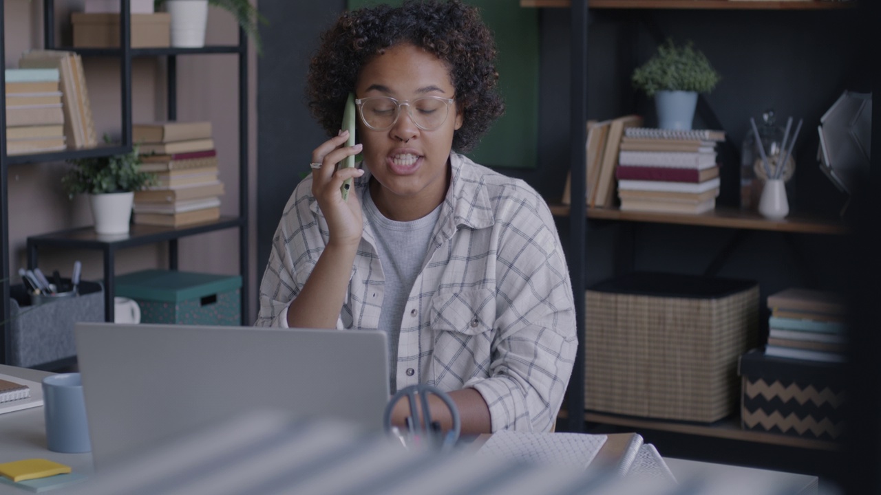 年轻的非裔美国女性自由职业者在家庭办公室用手机和笔记本电脑与客户交谈视频素材