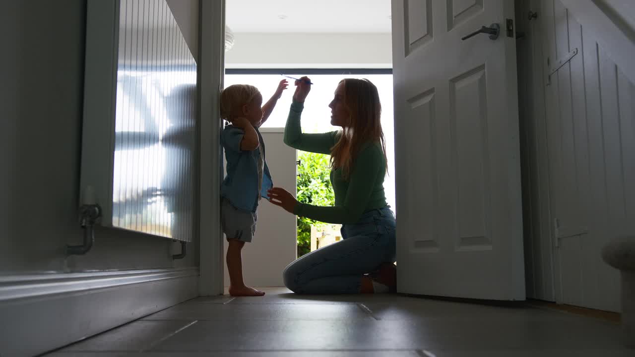 母亲在厨房里测量小儿子的身高，明亮的窗户映衬出他的剪影视频素材