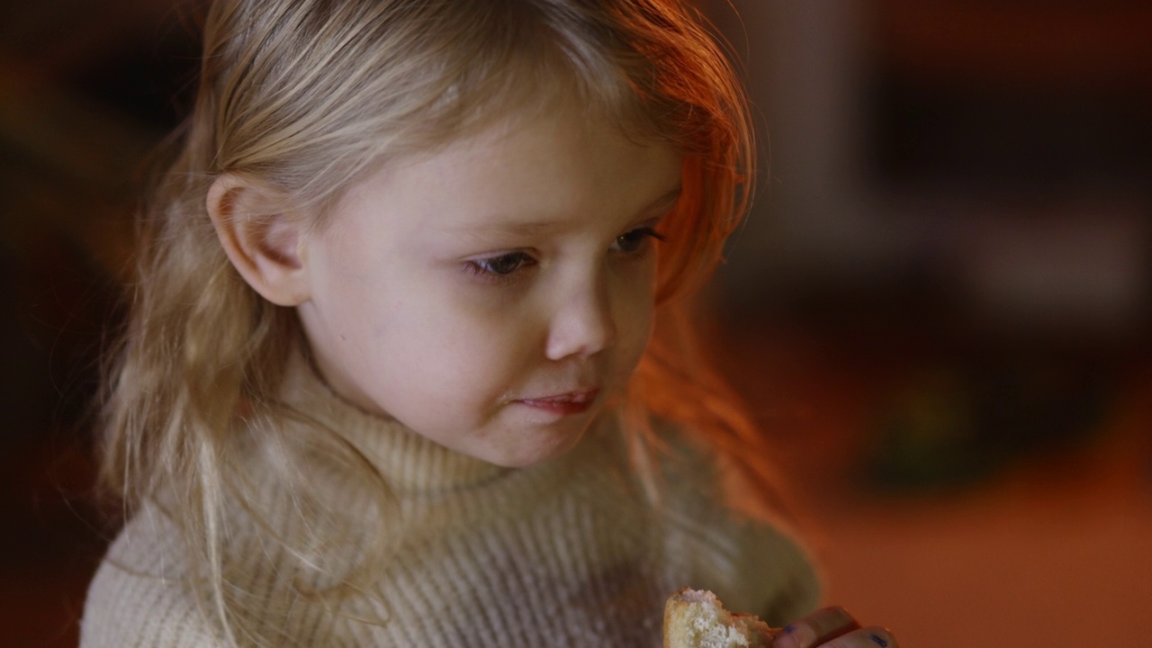 金发小女孩嚼着一块甜面包，温暖的衣服，蜡烛火的背景视频下载