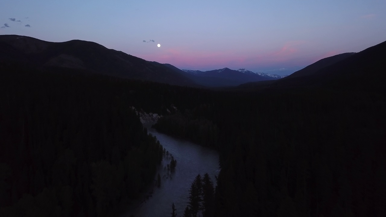 空中拍摄的河流在剪影山脉附近的树木中，无人机在黄昏时在自然景观上对着天空向后飞行-蒙大拿利比视频素材