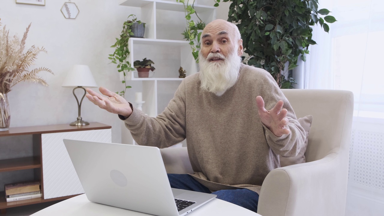 大胡子老人坐在笔记本电脑旁边的软扶手椅上对着镜头说话视频下载