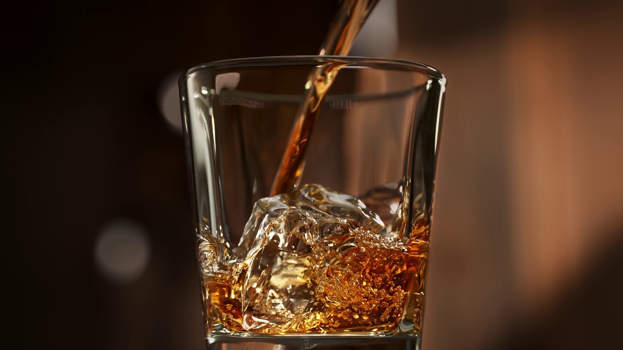 把威士忌倒进玻璃杯的冰块上，金色的液体在温暖的木制背景上以慢动作混合视频下载