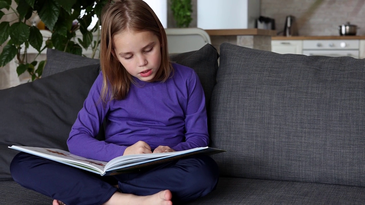 一个可爱的金发小女孩正坐在客厅的沙发上读一本彩色的大书。低龄学童自学，在家阅读文学作品，趣味休闲视频下载