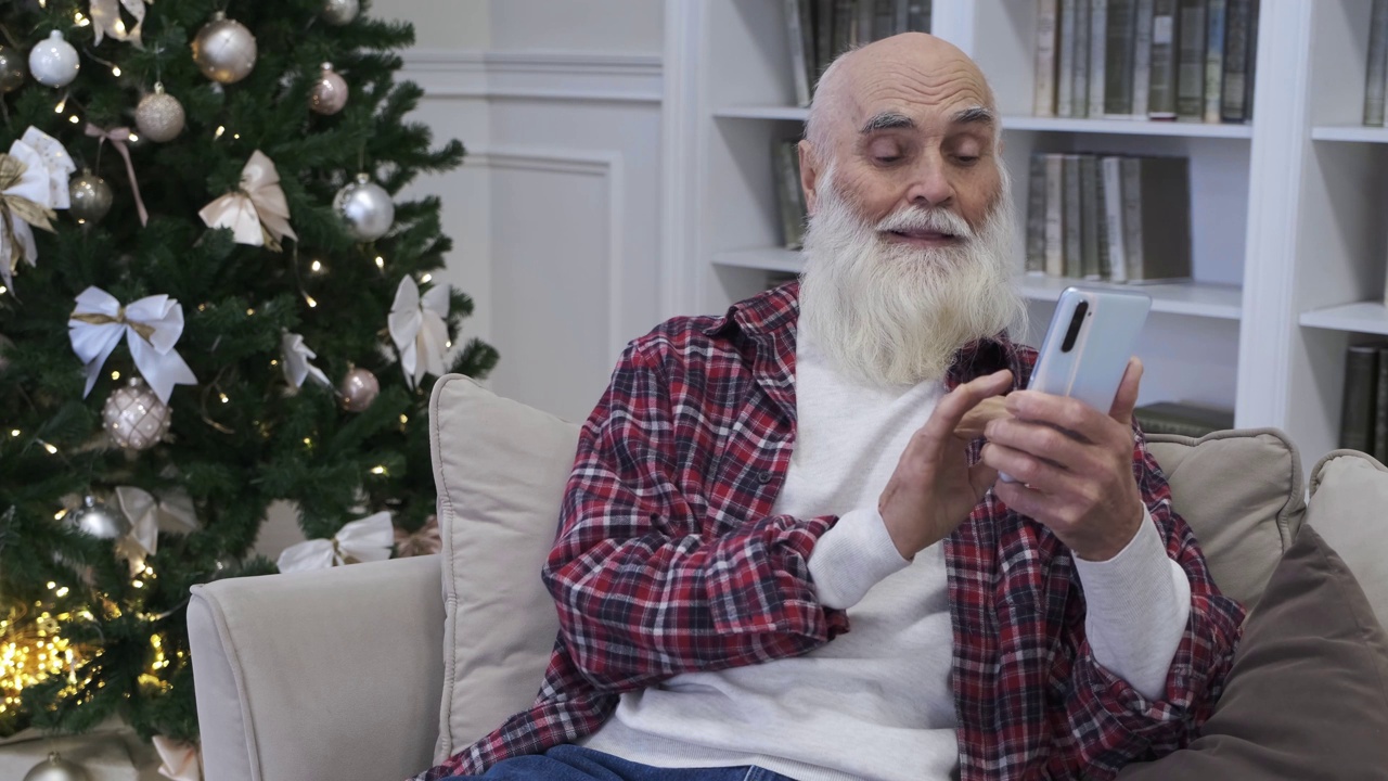 大胡子老人坐在舒适的沙发上看智能手机新闻视频下载