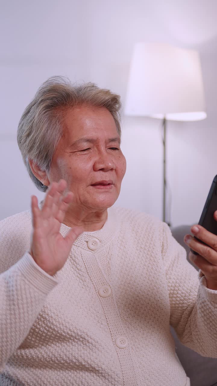 垂直夹。快乐的亚洲老太太坐在家里的沙发上玩智能手机。视频下载