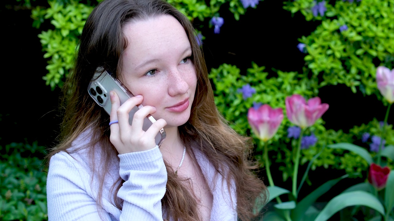 一个平静的交谈在电话上在街上一个年轻的女孩站在公园的灌木丛附近郁金香和蓝色的花上的药水说话扬起眉毛戳她的额头美丽的真实的人视频下载
