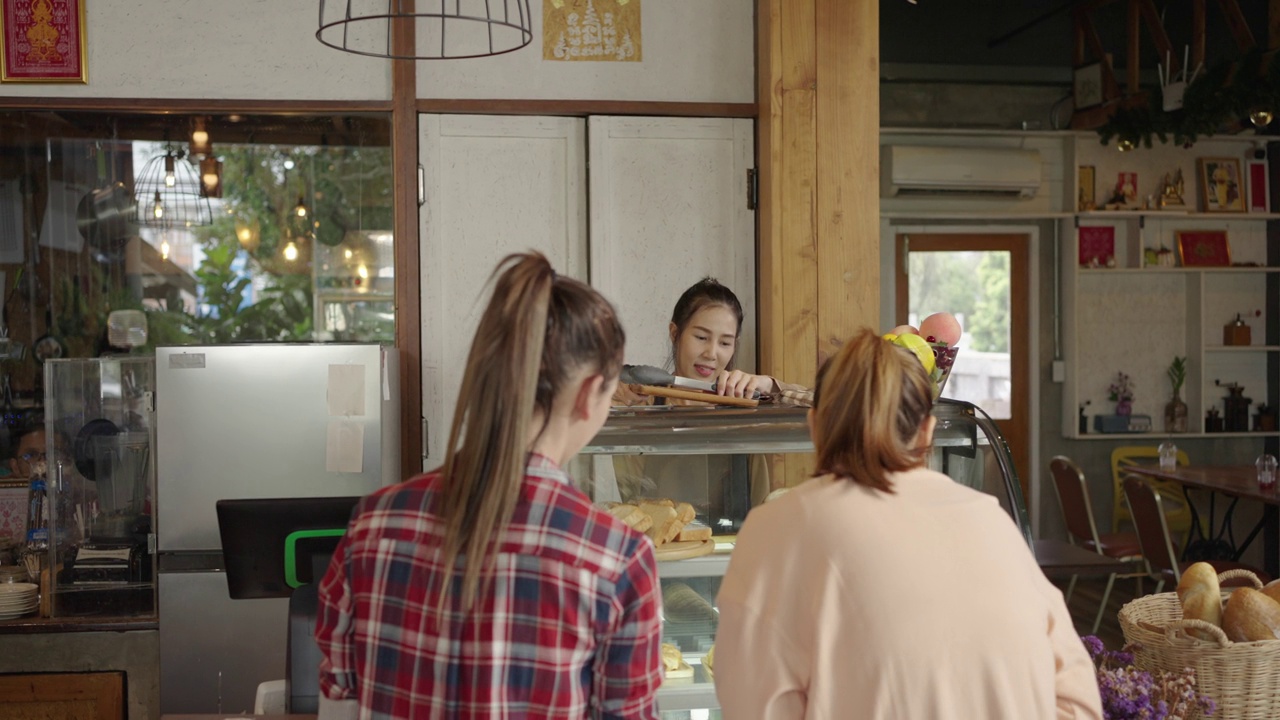 两个年轻女子在咖啡馆里从一个漂亮的亚洲女售货员那里挑选甜点或蛋糕。小企业，企业主。视频素材