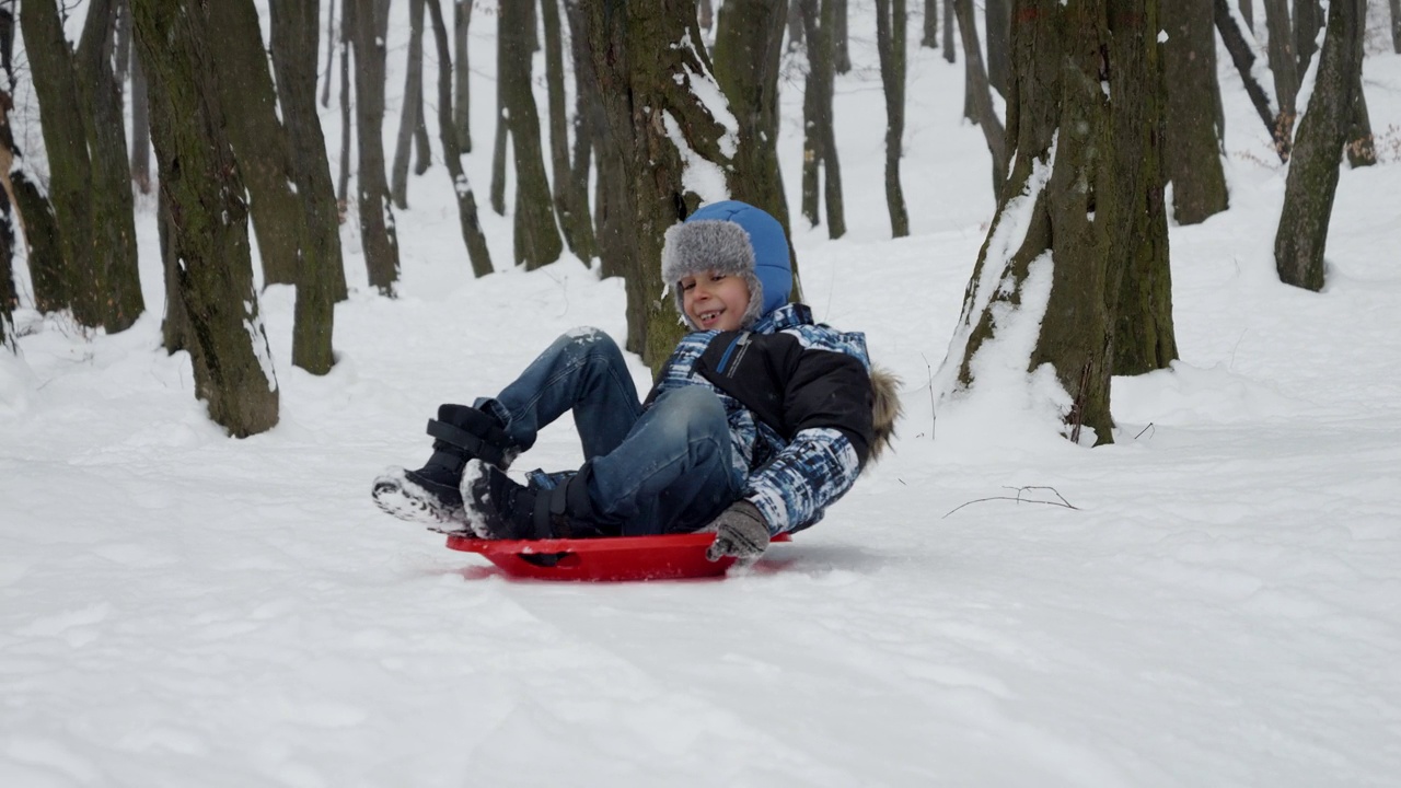 一个快乐微笑的小男孩骑着塑料雪橇从雪山上滑下的慢动作镜头。冬日假期的精髓和户外雪中玩耍的乐趣和兴奋视频素材