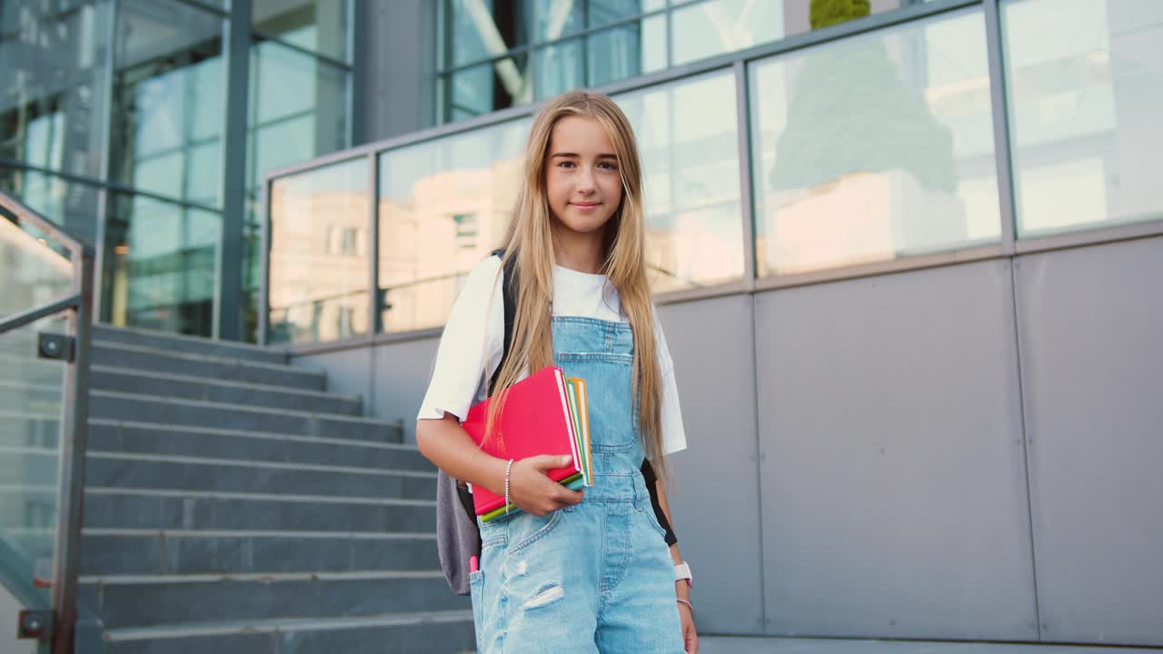 可爱的十几岁的女学生背着背包，在校园附近的相机前微笑着摆姿势。一名小学生手持课本站在学校大楼外的楼梯上。视频下载