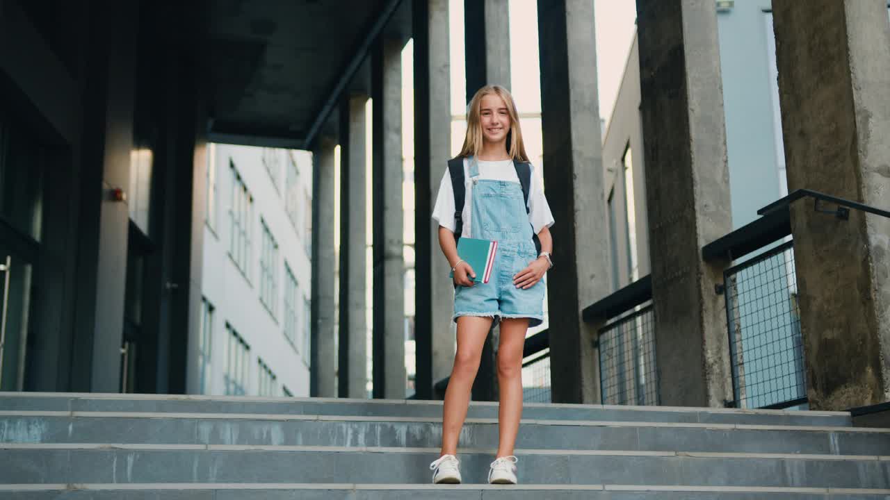 欢快的女孩抱着书，背着背包在学校大楼外摆姿势，对着镜头微笑。现代教育，大学学费和助学金，留学生和留学的概念。视频下载