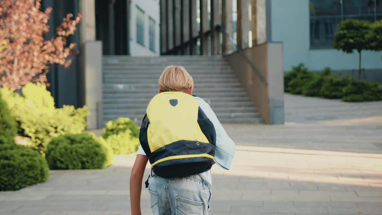 一个男生背着背包独自跑到太阳街的现代教学楼外面。一个小学生沿着城市街道的人行道跑去上学。儿童教育学习理念。视频下载