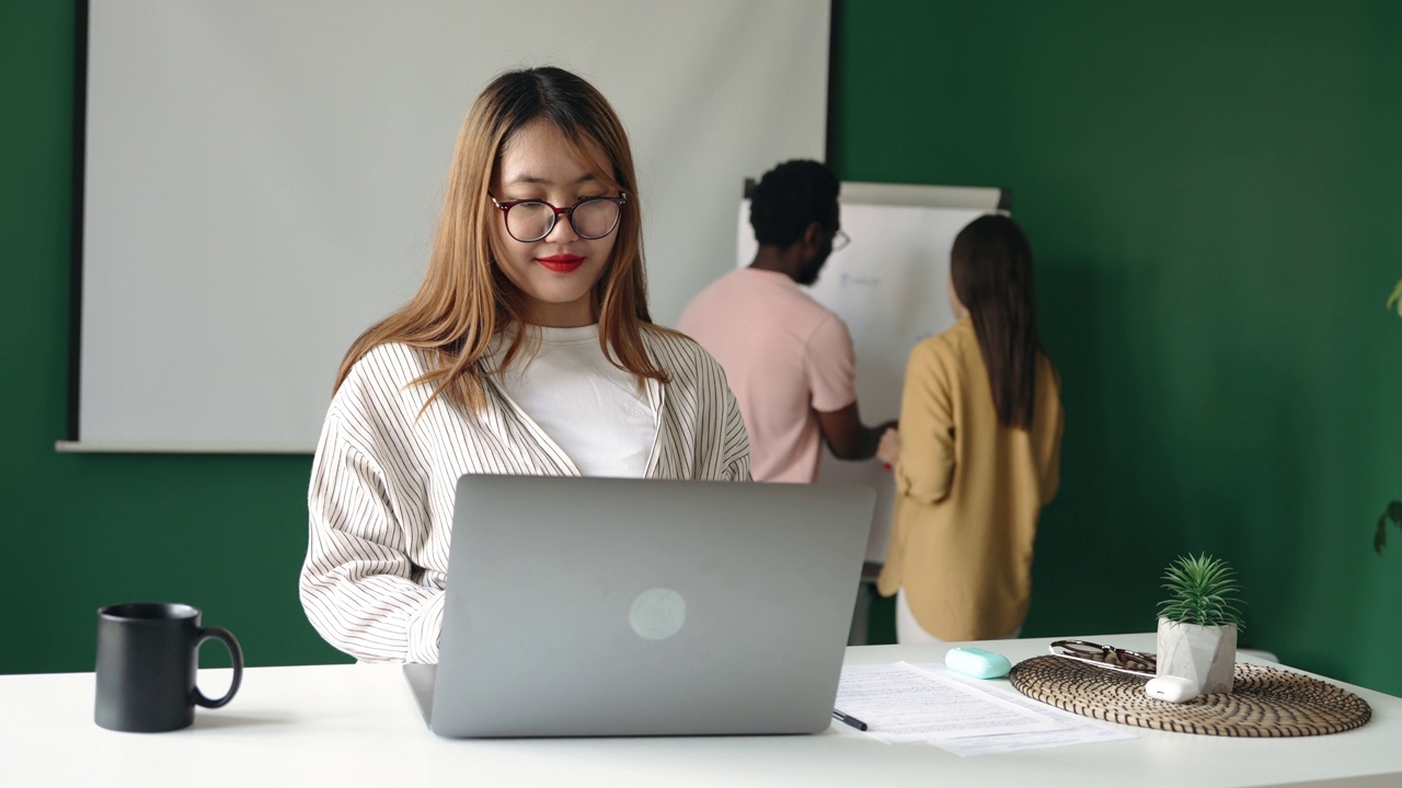 年轻的微笑的女人在现代化的办公室工作，使用笔记本电脑视频下载