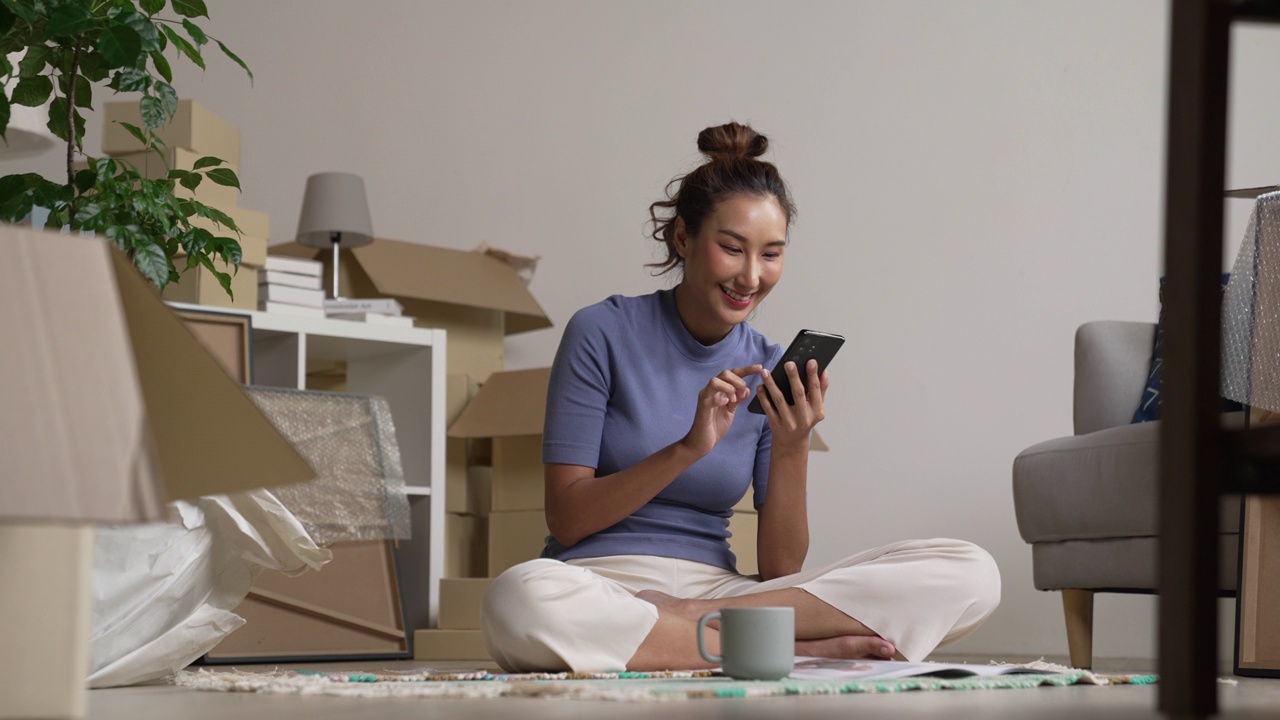快乐的年轻亚洲女性搬到新家，坐在地板上玩智能手机。视频下载
