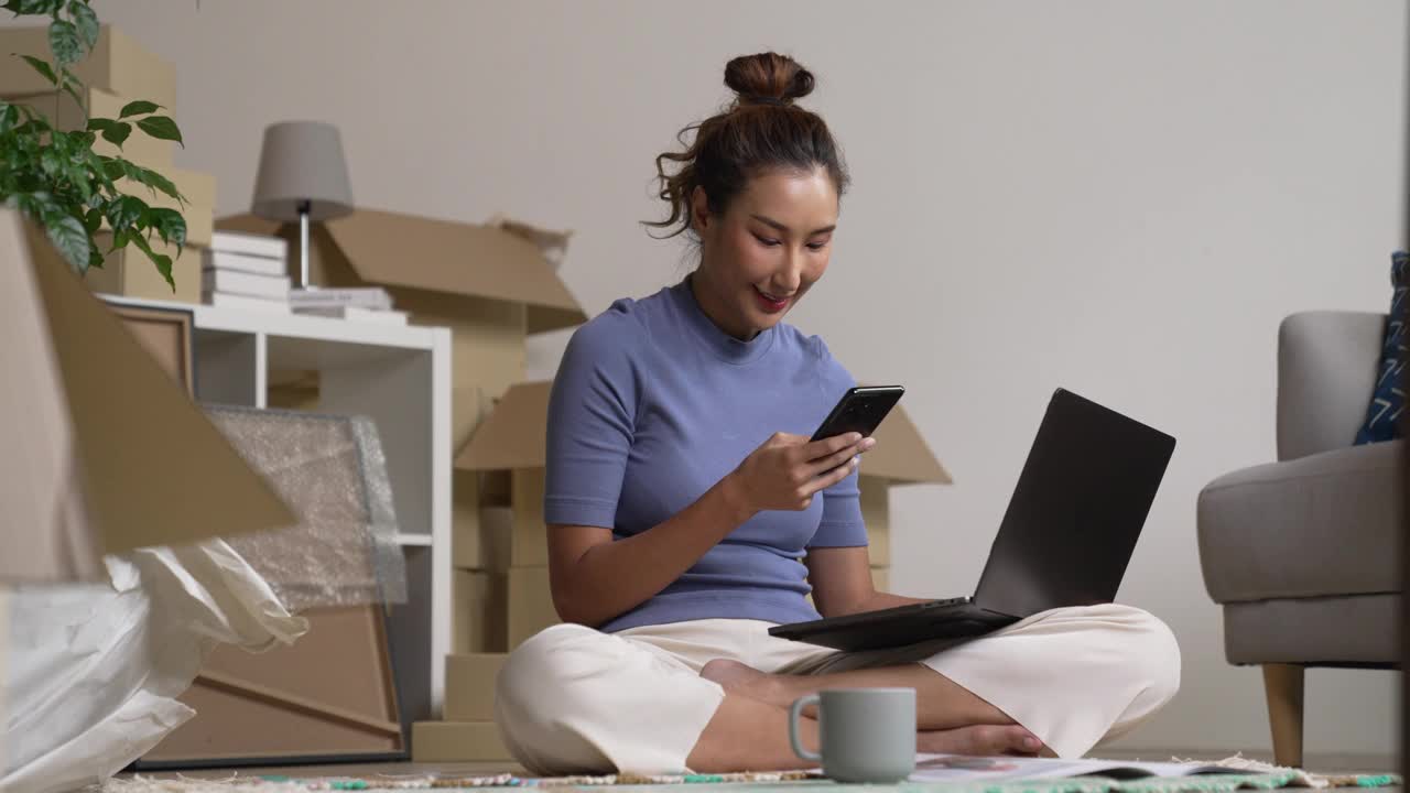 快乐的年轻亚洲女性搬到新家，坐在地板上使用智能手机和笔记本电脑。视频下载