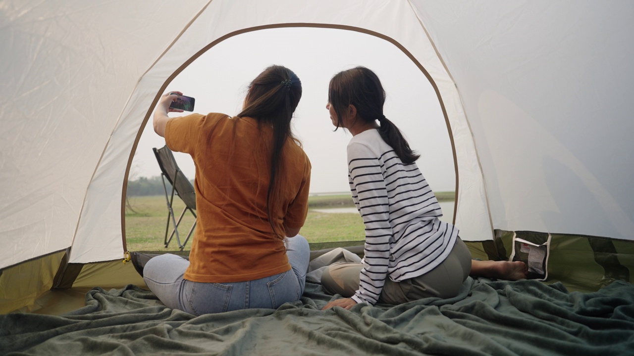 两个亚洲女孩坐在帐篷里用智能手机自拍，一起在户外露营视频下载