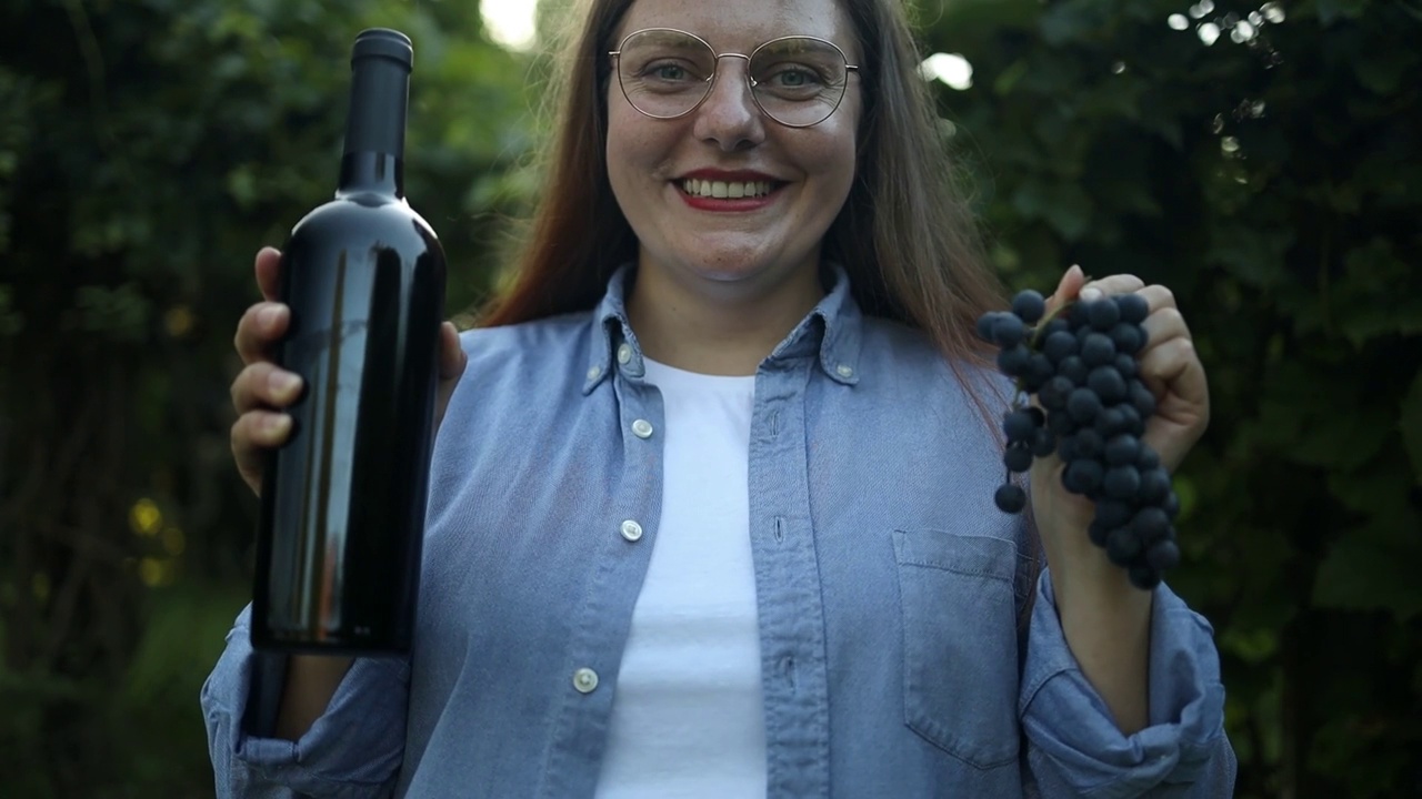 迷人的女农民的肖像展示葡萄和一瓶红葡萄酒生产在收获季节在日落的葡萄园可爱。酿酒葡萄收获。女侍酒师在品尝葡萄酒。视频素材