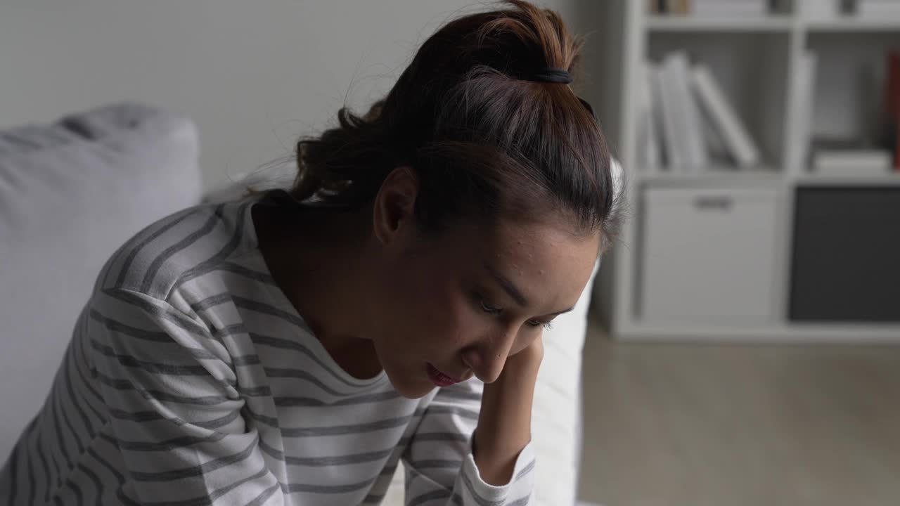 年轻的亚洲女性坐在家里的沙发上，感到悲伤、疲倦和担忧，心理健康状况堪忧。视频下载