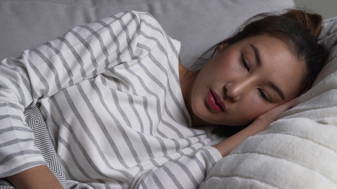 年轻的亚洲女性躺在家里的沙发上玩智能手机，感到悲伤、疲倦和担心，心理健康状况堪忧。视频下载