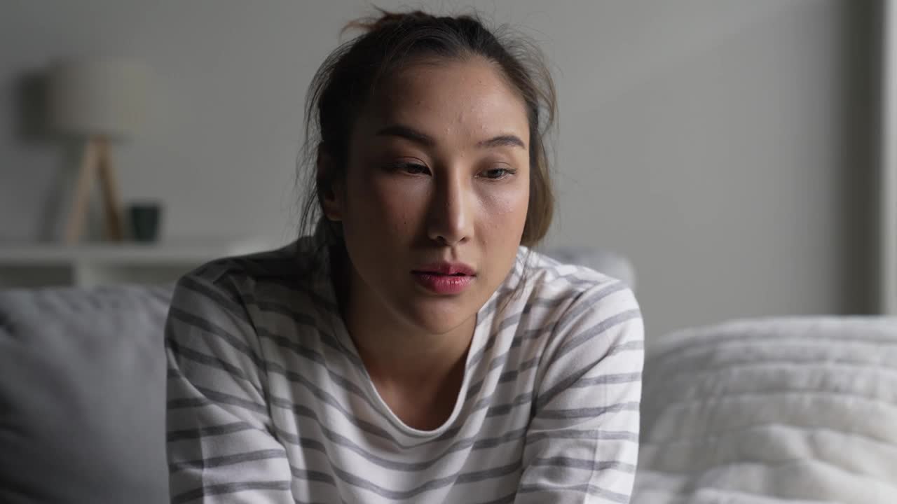 年轻的亚洲女性坐在家里的沙发上，感到悲伤、疲倦和担忧，心理健康状况堪忧。视频下载