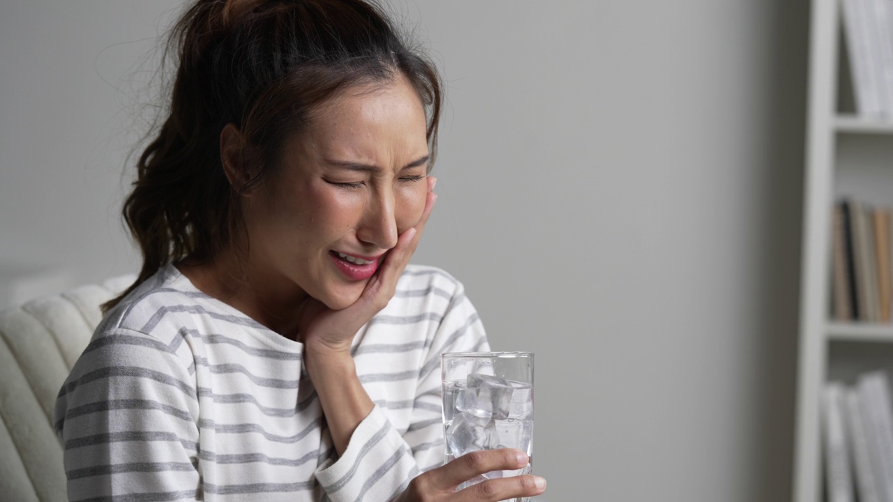 年轻的亚洲女子在喝装有冰块的水时牙痛。医疗保健理念。视频下载