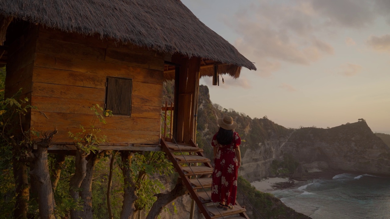 一个年轻的女游客站在传统的树屋，看着日出时的阿顿海滩，努沙佩尼达岛。印尼巴厘岛的热门旅游目的地。视频素材