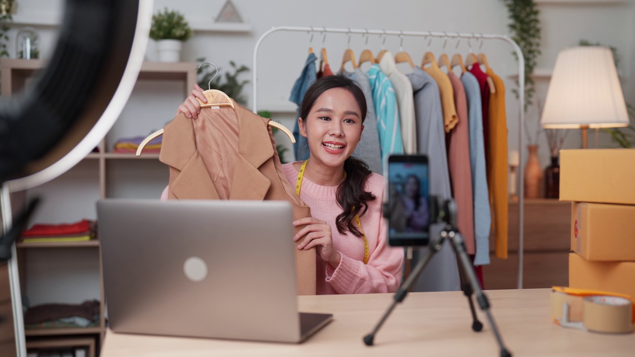 一名亚洲年轻成年女性正坐在办公桌前，通过智能手机直播销售自己的服装。视频下载